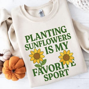 Pode incluir: Uma camisola bege de gola redonda com o texto verde "PLANTING SUNFLOWERS IS MY FAVORITE SPORT" e duas ilustrações de girassóis amarelos. A camisola é apresentada ao lado de três pequenas abóboras de tecido em laranja e bege.