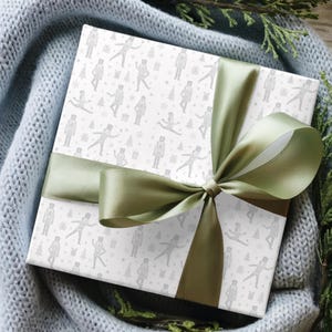 Puede incluir: Una caja de regalo envuelta con un fondo blanco y un patrón de cascanueces grises, copos de nieve y árboles de Navidad. La caja está atada con una gran cinta y lazo de satén verde oliva. El regalo descansa sobre una manta de punto gris.