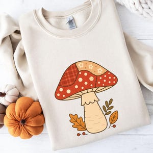 Puede incluir: Sudadera color crema con un diseño de hongo grande y caprichoso. El hongo tiene una gorra de retazos roja y naranja con puntos blancos, un tallo beige y hojas de otoño. Una prenda de vestir con temática otoñal.