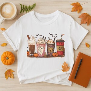 以下が含まれることがあります： ハロウィーンをテーマにした4つの飲み物のカラフルなイラストが描かれた白いTシャツ。各飲み物は、幽霊、コウモリ、カボチャなどの不気味な要素で飾られています。飲み物の色は、茶色、ピンク、赤など様々です。