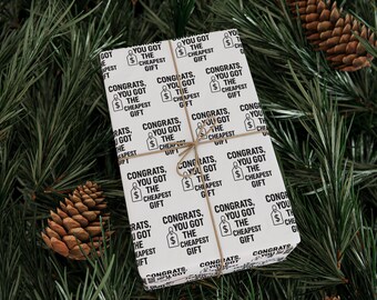 Könnte beinhalten: Ein verpacktes Geschenk mit weißem Hintergrund und schwarzem Text mit der Aufschrift "Herzlichen Glückwunsch, Sie haben das billigste Geschenk bekommen". Das Geschenk ist mit Bindfaden gebunden und liegt auf einem Bett aus grünen Tannennadeln und Tannenzapfen.
