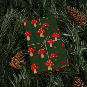 **Alt Text:**
Gift wrapped in festive mushroom-themed Christmas paper featuring red toadstools, holly berries, and golden stars on a green background, tied with twine and placed among pine branches and pinecones.