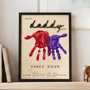 Può includere: Un cartello in legno incorniciato con le parole "BEST daddy EVER" in corsivo nero. Il cartello presenta impronte di mani rosse e viola e il testo "HANDS DOWN LOVE, Olive & Olivia". Il cartello è esposto su una mensola di legno.