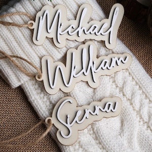 May include: Three wooden name tags with the names "Michael", "William", and "Sienna" in a layered design. Each tag has a white script font on a light wood base and is attached to a jute string. The tags are displayed on a white cable-knit surface.
