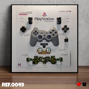 May include: A framed display of a PlayStation DualShock controller, deconstructed to show its components. Labels such as "L1", "R2", and "Buttons" are present. The PlayStation logo is displayed. The text "REF.0049" is at the bottom of the frame.