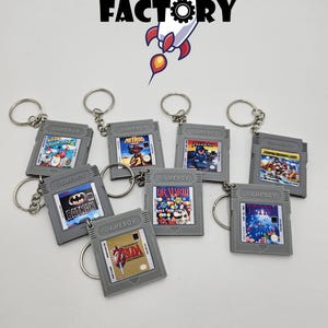 Può includere: Una collezione di portachiavi a forma di cartuccia Gameboy grigie, con titoli di giochi come "Batman", "Dr. Mario" e "Zelda". I portachiavi sono disposti su una superficie bianca, con il logo "MAD FACTORY" e una grafica a razzo sullo sfondo.