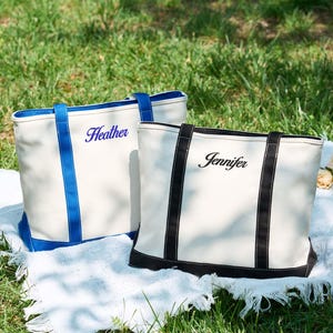 May include: Two white canvas tote bags with personalized names, "Heather" in blue and "Jennifer" in black script. The bags have contrasting trim and handles, with one in blue and the other in black. They are displayed on a white blanket on green grass.