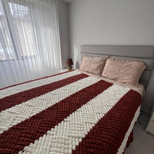 Könnte beinhalten: Eine rot-weiß gestreifte gestrickte Wolldecke auf einem Bett mit einem grauen Kopfteil. Die Decke ist aus dickem Garn gefertigt und hat eine gewebte Textur.