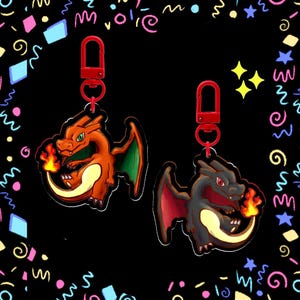May include: Two acrylic keychains featuring dragon-like creatures with flames on their tails. One creature is orange with green wings, and the other is dark gray with red wings. Both have red lobster claw clasps and are set against a black background.