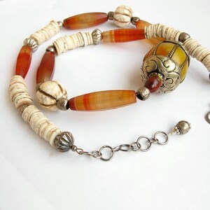 May include: A long necklace with a mix of white, orange, and brown beads. The necklace has a large, ornate, yellow and silver bead in the center. The necklace is finished with a silver clasp.