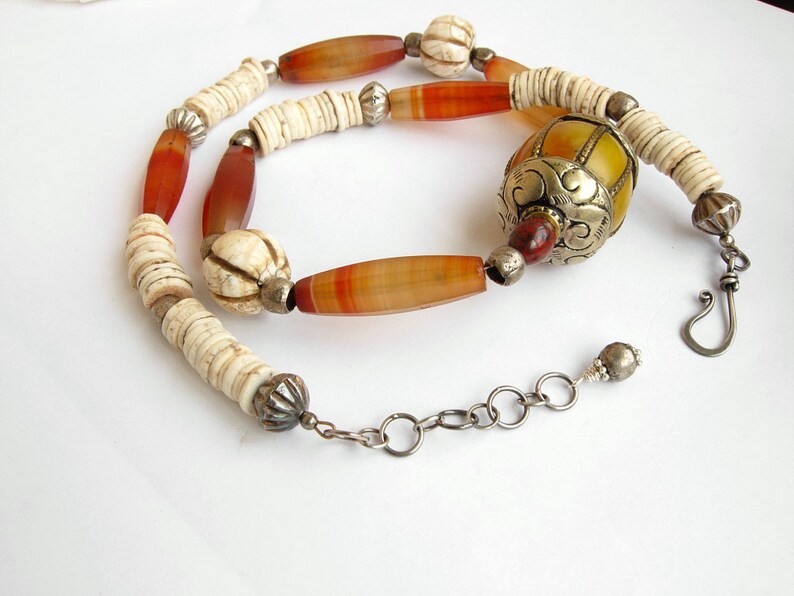 May include: A long necklace with a mix of white, orange, and brown beads. The necklace has a large, ornate, yellow and silver bead in the center. The necklace is finished with a silver clasp.