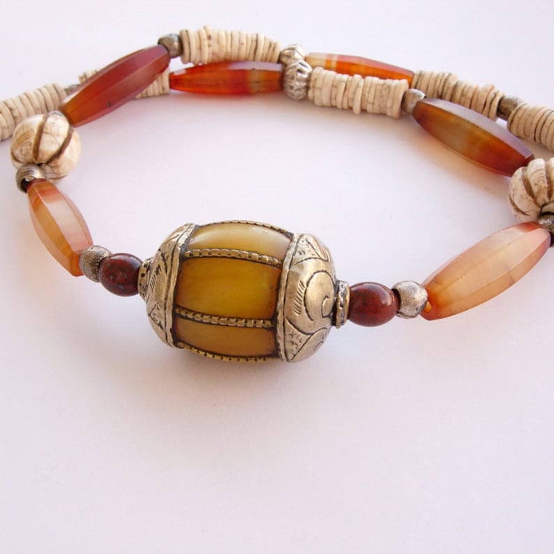 May include: A necklace made of brown, white, and red beads with a large, yellow, barrel-shaped bead in the center. The bead is decorated with silver accents.