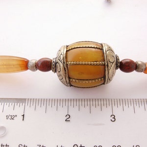 May include: A close-up of a necklace with a large amber bead surrounded by smaller carnelian beads and silver spacers. The amber bead is about 1 inch in diameter and has a decorative silver design. The necklace is measured against a ruler.