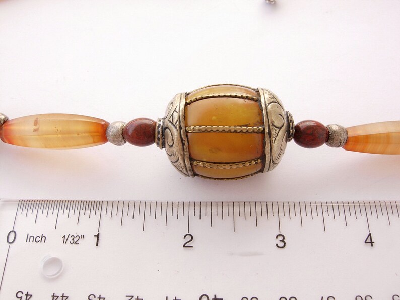 May include: A close-up of a necklace with a large amber bead surrounded by smaller carnelian beads and silver spacers. The amber bead is about 1 inch in diameter and has a decorative silver design. The necklace is measured against a ruler.
