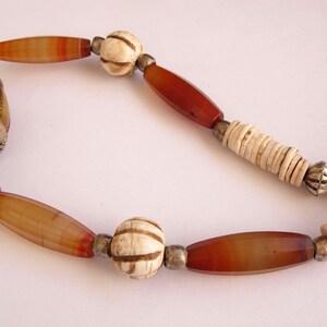 May include: A necklace made of carnelian, bone, and metal beads. The necklace features a large, yellow, carved bead with silver accents.