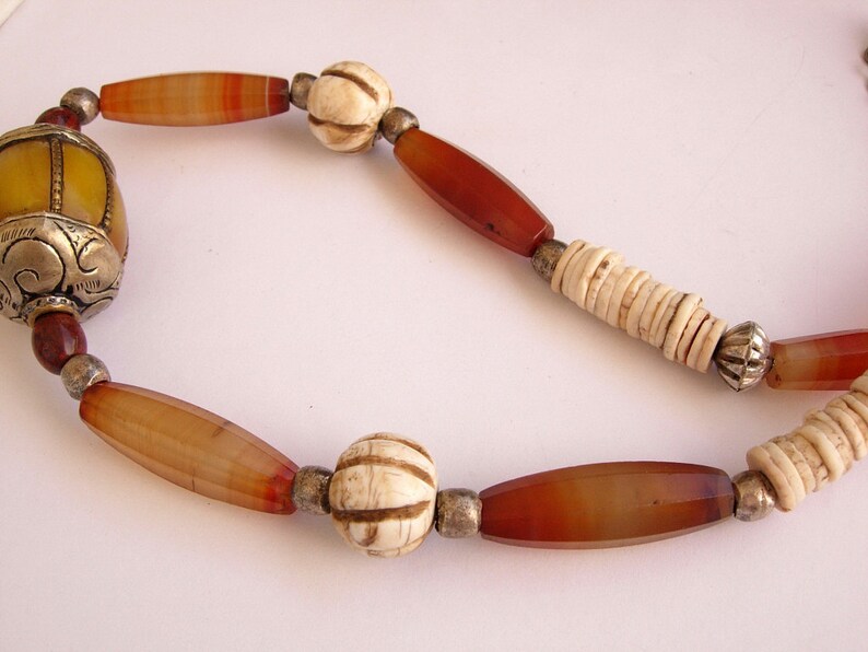 May include: A necklace made of carnelian, bone, and metal beads. The necklace features a large, yellow, carved bead with silver accents.