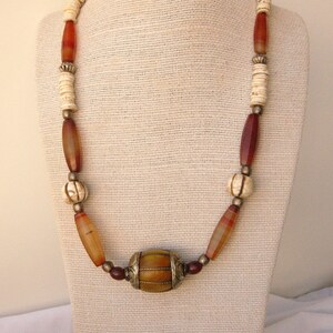May include: A long necklace made of brown, red, and white beads. The necklace has a large, round, gold-colored bead in the center.