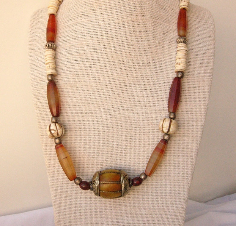 May include: A long necklace made of brown, red, and white beads. The necklace has a large, round, gold-colored bead in the center.