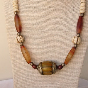 May include: A necklace made of brown, orange, and white beads with a large, round, yellow and brown bead in the center. The necklace is on a white fabric background.