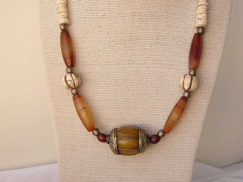May include: A necklace made of brown, orange, and white beads with a large, round, yellow and brown bead in the center. The necklace is on a white fabric background.