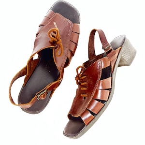 May include: A pair of brown leather sandals with open-toe design. The sandals feature a strap around the ankle and a small heel. The upper part of the sandals has a woven design with a lace-up detail. The sandals are set against a white background.