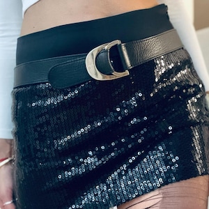 K&ouml;nnte beinhalten: Schwarzer Minirock mit Pailletten und einem schwarzen Lederg&uuml;rtel mit silberner Schnalle. Der Rock wird mit einem wei&szlig;en Langarm-Top und schwarzen Strumpfhosen kombiniert.
