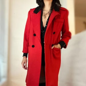 May include: A long, bright red coat with a black collar and black buttons. The coat has a double-breasted design and pockets. The sleeves are long, and the coat appears to be made of a wool or wool-blend material. The coat is knee-length.
