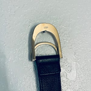 K&ouml;nnte beinhalten: Nahaufnahme eines schwarzen Lederg&uuml;rtels mit einer silberfarbenen Schnalle. Die Schnalle hat ein gebogenes Design mit einem kleinen, runden Schmuckelement. Der Markenname "VERSACE" ist auf der Schnalle eingraviert.