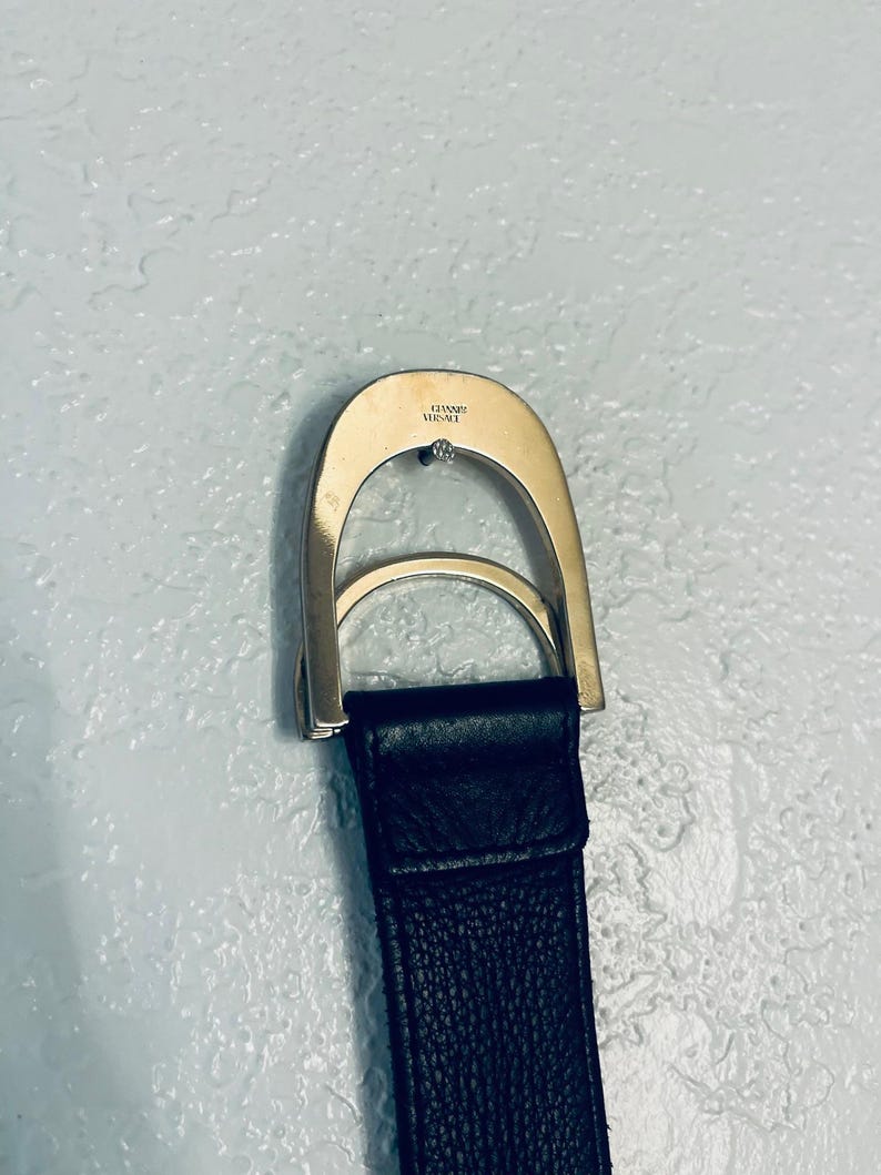 K&ouml;nnte beinhalten: Nahaufnahme eines schwarzen Lederg&uuml;rtels mit einer silberfarbenen Schnalle. Die Schnalle hat ein gebogenes Design mit einem kleinen, runden Schmuckelement. Der Markenname "VERSACE" ist auf der Schnalle eingraviert.