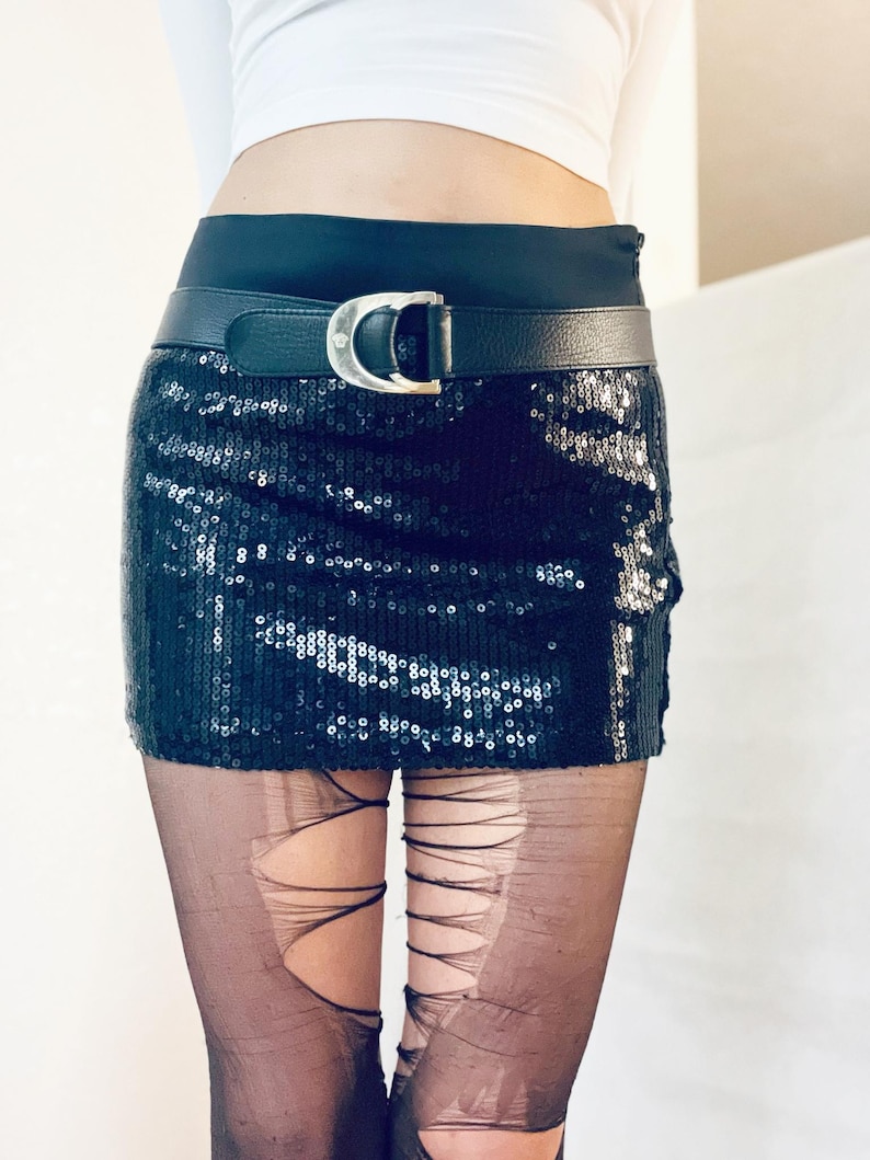 K&ouml;nnte beinhalten: Schwarzer Pailletten-Minirock mit schwarzem Lederg&uuml;rtel und silberner Schnalle. Der Rock wird mit zerrissenen schwarzen Strumpfhosen kombiniert. Ein wei&szlig;es Crop-Top vervollst&auml;ndigt den Look.