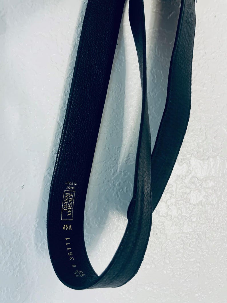 K&ouml;nnte beinhalten: Schwarzer Lederg&uuml;rtel mit dem Text "GIANNI VERSACE" und "MADE IN ITALY". Der G&uuml;rtel ist 75 cm lang. Der G&uuml;rtel ist dunkel und hat eine strukturierte Oberfl&auml;che.