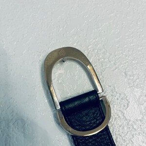 K&ouml;nnte beinhalten: Schwarzer Lederg&uuml;rtel mit silberfarbener Schnalle. Die Schnalle hat ein D-Ring-Design und ein kleines Emblem. Der G&uuml;rtel hat eine strukturierte Oberfl&auml;che.