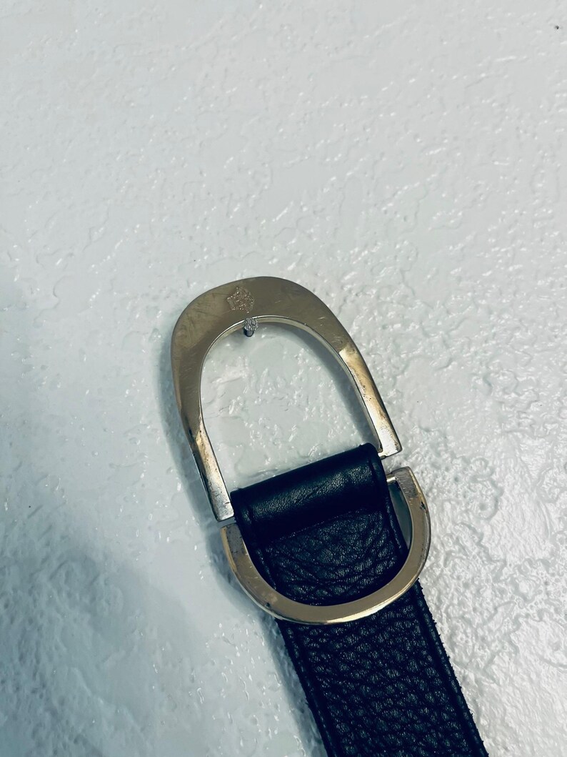 K&ouml;nnte beinhalten: Schwarzer Lederg&uuml;rtel mit silberfarbener Schnalle. Die Schnalle hat ein D-Ring-Design und ein kleines Emblem. Der G&uuml;rtel hat eine strukturierte Oberfl&auml;che.