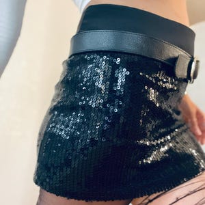K&ouml;nnte beinhalten: Schwarzer Minirock mit Pailletten, schwarzem G&uuml;rtel und silberner Schnalle. Der Rock besteht aus kleinen, gl&auml;nzenden Pailletten, die das Licht reflektieren. Der G&uuml;rtel ist aus schwarzem Leder. Getragen mit schwarzer Strumpfhose und wei&szlig;em Oberteil.