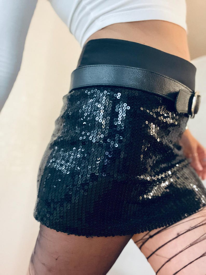 K&ouml;nnte beinhalten: Schwarzer Minirock mit Pailletten, schwarzem G&uuml;rtel und silberner Schnalle. Der Rock besteht aus kleinen, gl&auml;nzenden Pailletten, die das Licht reflektieren. Der G&uuml;rtel ist aus schwarzem Leder. Getragen mit schwarzer Strumpfhose und wei&szlig;em Oberteil.