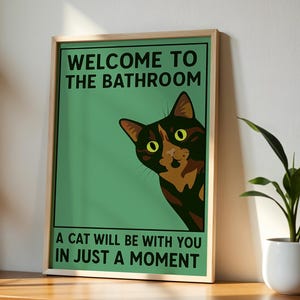 Op de afbeelding: Ingelijste poster met een groene achtergrond en de tekst "WELCOME TO THE BATHROOM" en "A CAT WILL BE WITH YOU IN JUST A MOMENT." Een kat met gele ogen piekt van de rechterkant van de poster. De lijst is lichtbruin.