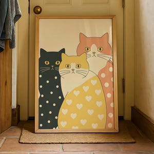 May include: A framed art print featuring three stylized cats. One cat is black with white polka dots, another is yellow with white hearts, and the third is peach with pink polka dots. The artwork is displayed in a light-colored wooden frame.