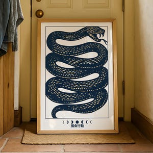 Pode incluir: Uma impressão artística emoldurada com uma ilustração azul marinho de uma cobra enrolada em um fundo branco. A obra de arte inclui texto japonês e símbolos de fases da lua. A moldura é marrom claro.