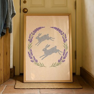 May include: A framed print showing two grey hares leaping within a lavender wreath. The artwork has a light beige background and is displayed in a natural wood frame. The print is leaning against a door.