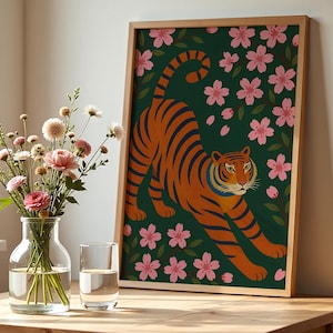 May include: A framed art print featuring an orange tiger with blue stripes, set against a dark green background with pink cherry blossoms. The artwork is displayed on a wooden table, next to a vase of flowers and a glass of water.