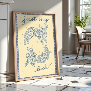 May include: A framed art print featuring two blue rabbit illustrations with floral patterns on a light yellow background. The words "just my luck" are written in blue script. The print has a light wood frame and a blue border.