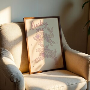 May include: A framed art print featuring two stylized rabbits with floral patterns in a soft pink hue. The words "lucky me" are written in cursive at the top. The artwork is displayed on a beige armchair.