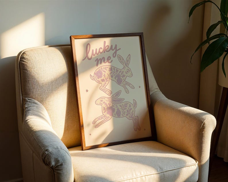 May include: A framed art print featuring two stylized rabbits with floral patterns in a soft pink hue. The words "lucky me" are written in cursive at the top. The artwork is displayed on a beige armchair.