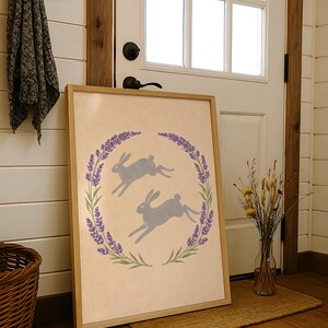 May include: A framed print showing two grey hares leaping inside a lavender and green wreath. The artwork has a light beige background and a natural wood frame. The print is leaning against a white door.