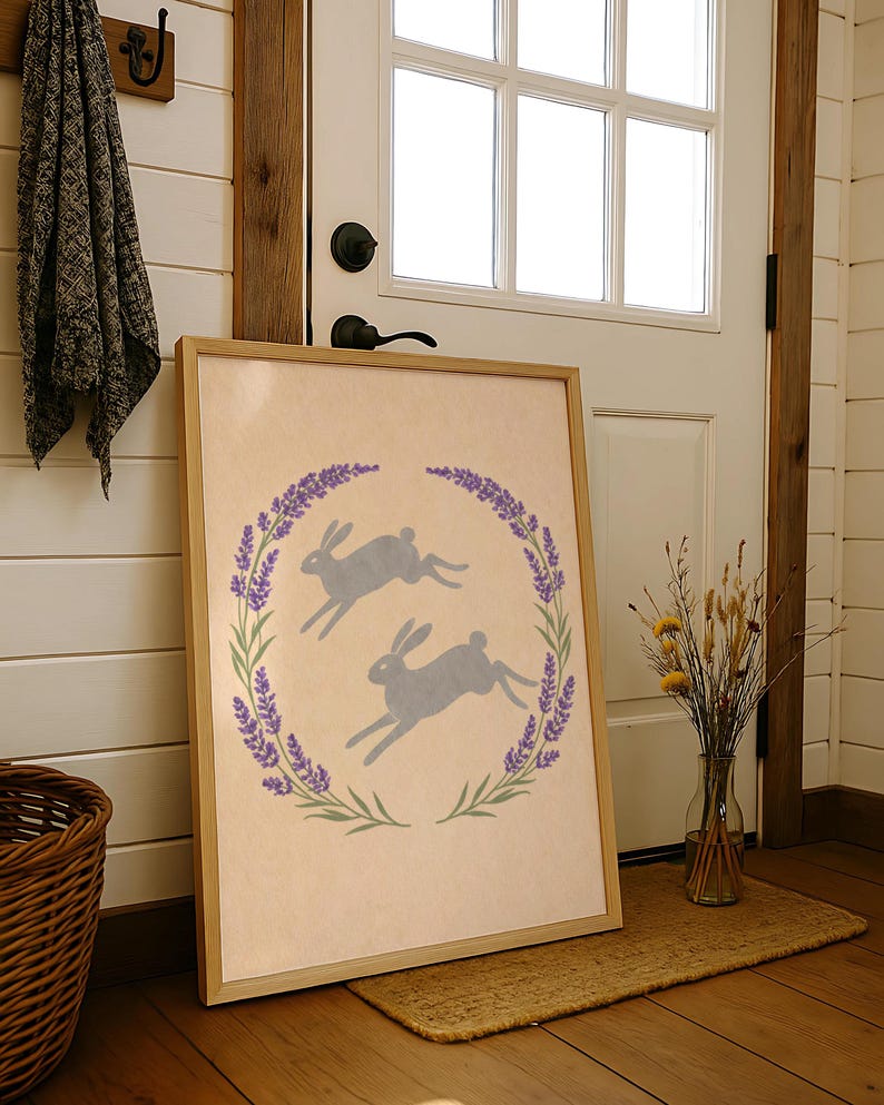 May include: A framed print showing two grey hares leaping inside a lavender and green wreath. The artwork has a light beige background and a natural wood frame. The print is leaning against a white door.