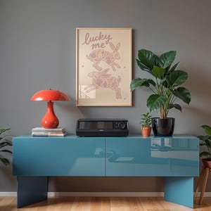 May include: A teal blue cabinet with a red lamp, a black appliance, and a framed print that says "lucky me" with rabbit illustrations. Potted plants are on the cabinet and on the floor.