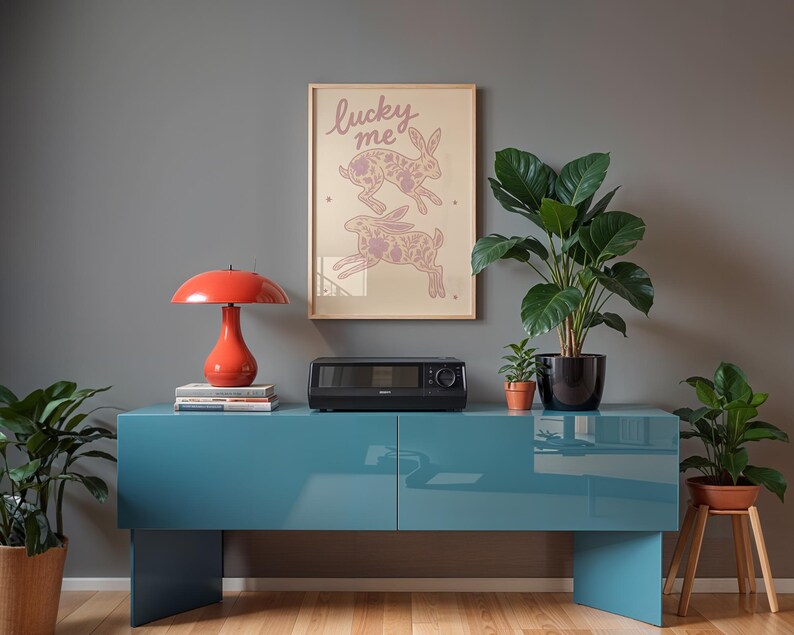 May include: A teal blue cabinet with a red lamp, a black appliance, and a framed print that says "lucky me" with rabbit illustrations. Potted plants are on the cabinet and on the floor.