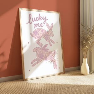 May include: A framed art print featuring two running rabbits with floral patterns in shades of pink. The words "lucky me" are written in cursive at the top. The print is set against a white background with small star accents.