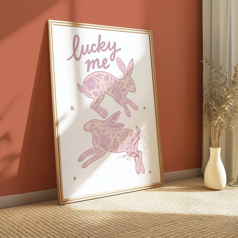 May include: A framed art print featuring two running rabbits with floral patterns in shades of pink. The words "lucky me" are written in cursive at the top. The print is set against a white background with small star accents.