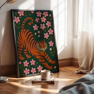 May include: Framed art print featuring an orange tiger with black stripes, stretching amidst pink cherry blossoms on a dark green background. The artwork is in a black frame and leans against a wall. A coffee cup and book are in front of the artwork.